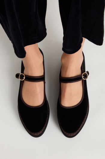 A person is wearing Black Velvet Platform Mary Jane Shoes with buckle straps, standing on a light-colored surface. The handmade shoes are styled with dark pants.