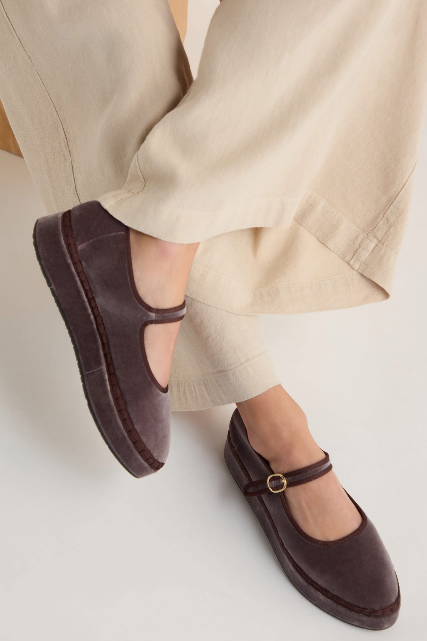A person in beige pants and Taupe Velvet Platform Mary Jane Shoes, handmade in Spain with a cotton velvet upper and buckle strap, sits cross-legged.