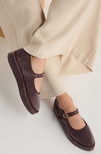 A person in beige pants and Taupe Velvet Platform Mary Jane Shoes, handmade in Spain with a cotton velvet upper and buckle strap, sits cross-legged.