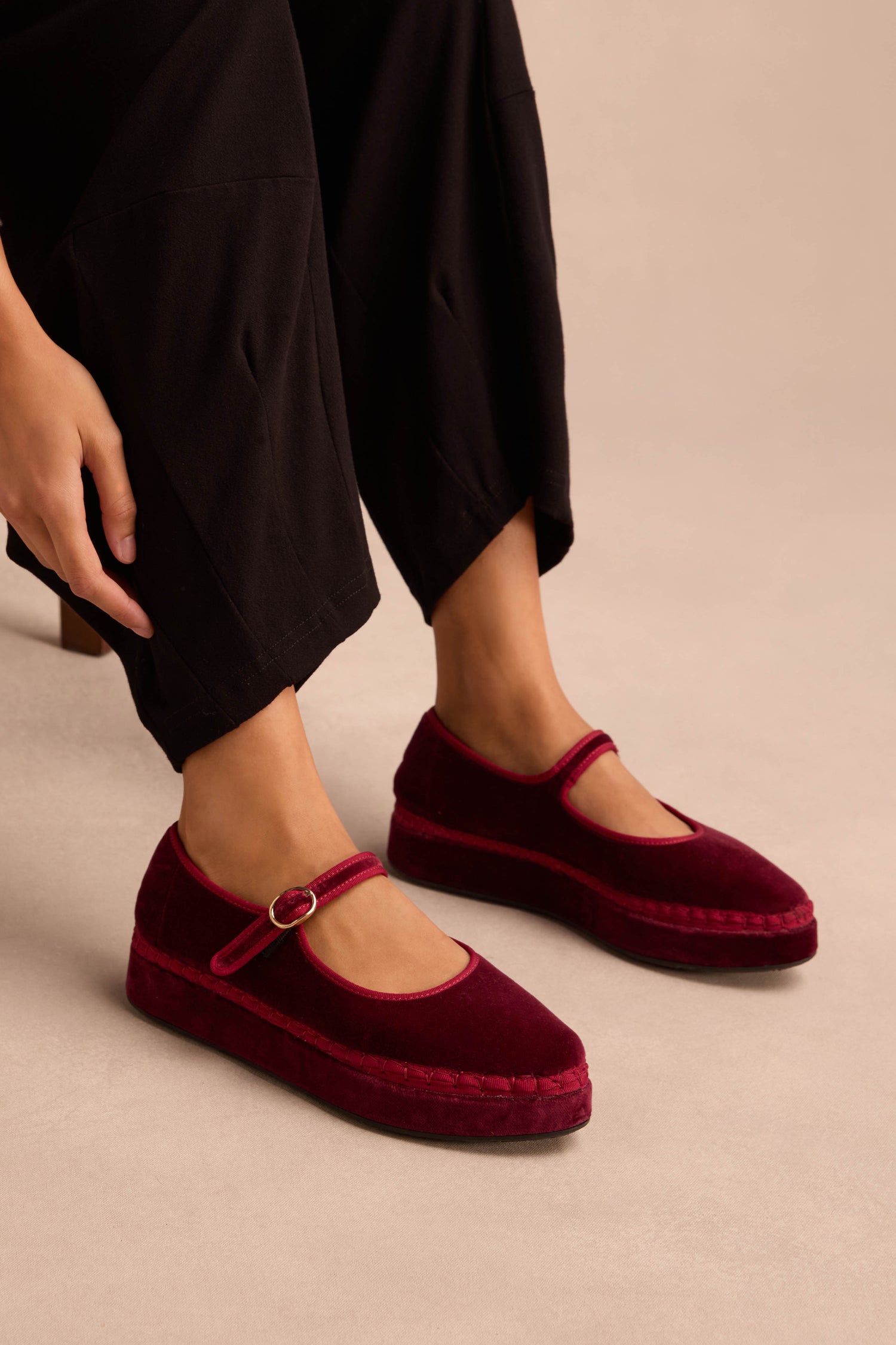 A person wearing black pants and Wine Velvet Platform Mary Jane Shoes with buckle straps, handmade in Spain, is seated with one hand resting near their ankle.
