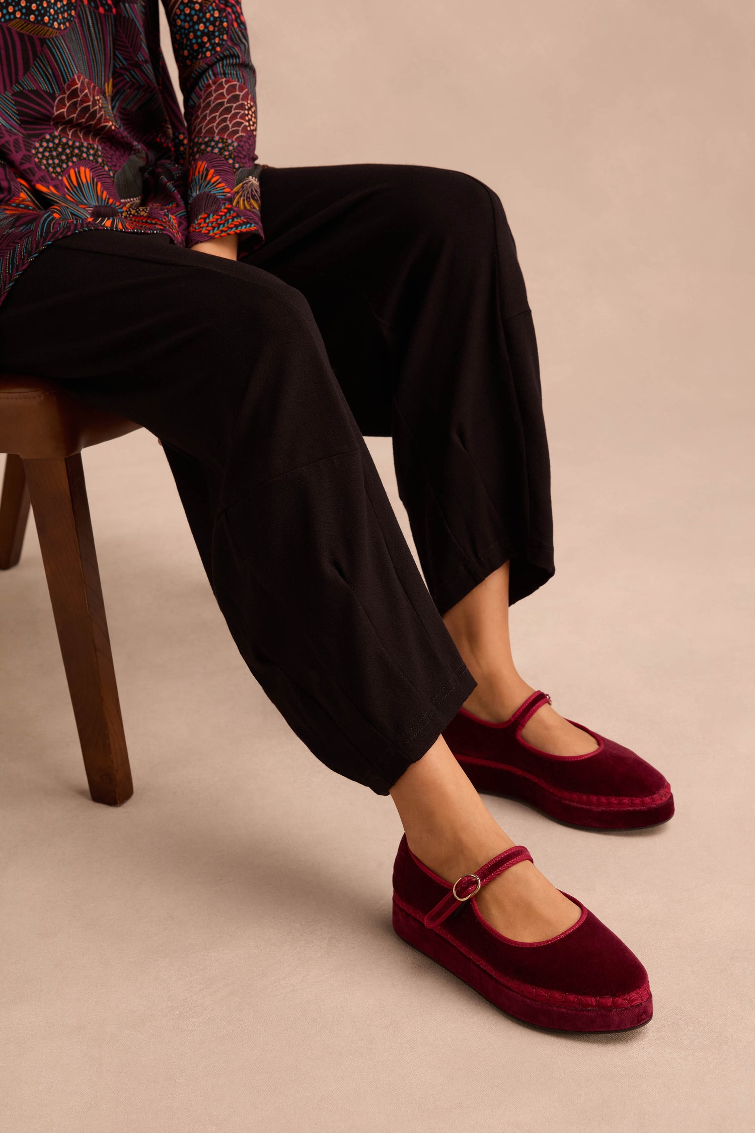 A person seated on a chair wears black pants and eye-catching Wine Velvet Platform Mary Jane Shoes with ankle straps, handmade in Spain. Only the lower half of their outfit is visible against a neutral background.