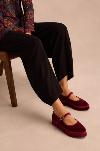 A person seated on a chair wears black pants and eye-catching Wine Velvet Platform Mary Jane Shoes with ankle straps, handmade in Spain. Only the lower half of their outfit is visible against a neutral background.