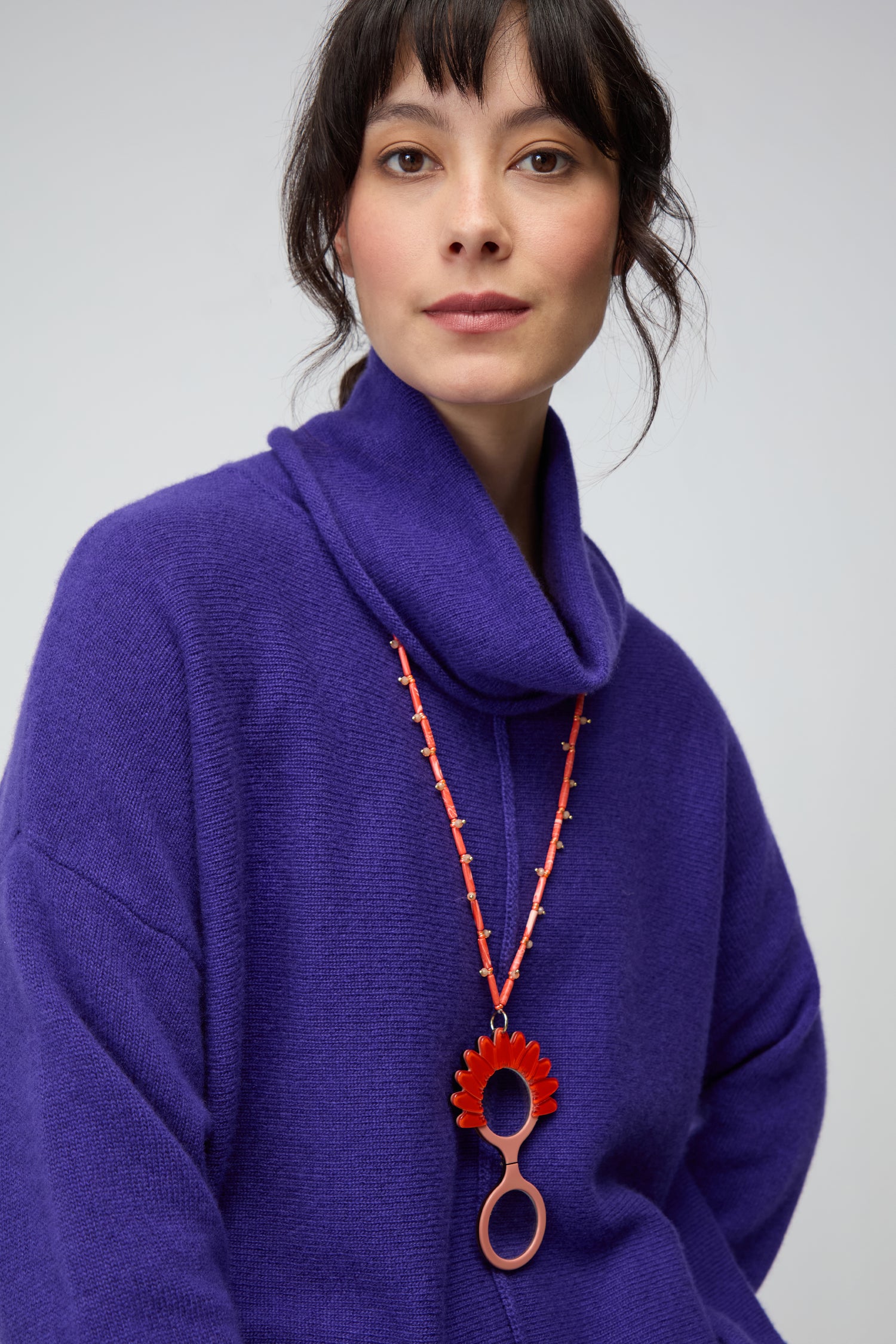A woman in a vibrant purple turtleneck and a chunky red necklace with floral and looped accents, inspired by the Daisy Pendant Glasses design, looks at the camera against a plain background.