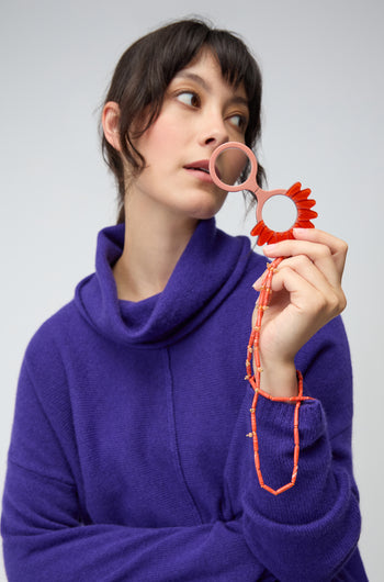 A woman in a purple sweater holds the Daisy Pendant Glasses, featuring a round mirror with a red flower frame and beaded handle, as she looks thoughtfully to the side.