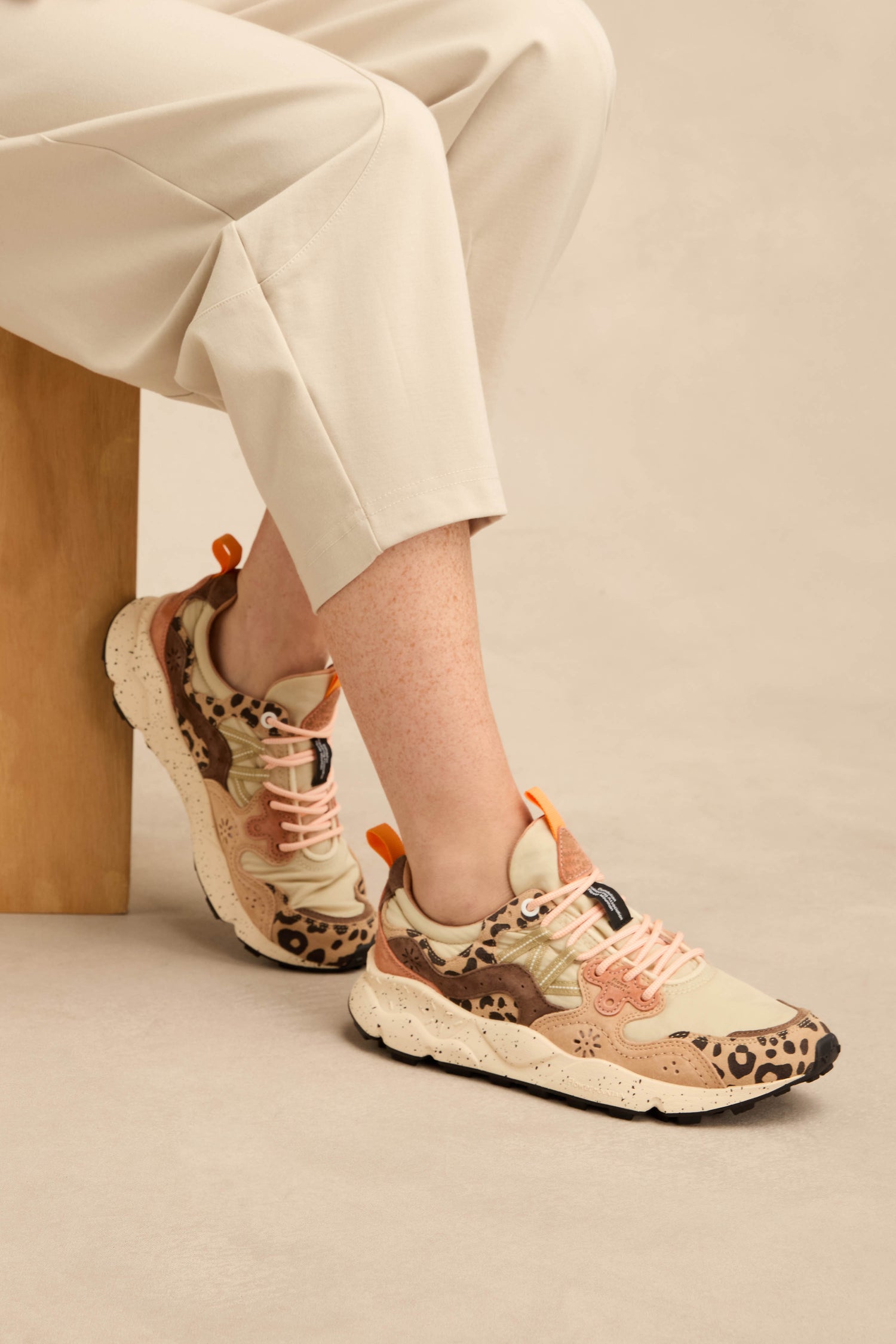 A person seated on a light wooden bench against a neutral background wears beige pants and Yamano 3 Cheetah Detail Trainers featuring suede uppers, pink laces, and cheetah-patterned accents.