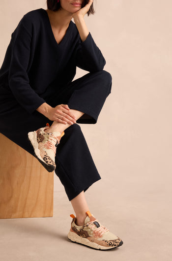 A person seated on a light wooden bench against a neutral background wears beige pants and Yamano 3 Cheetah Detail Trainers featuring suede uppers, pink laces, and cheetah-patterned accents.