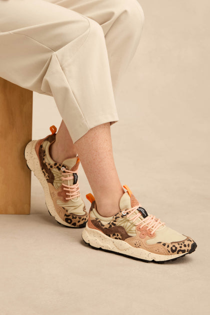 A person seated on a light wooden bench against a neutral background wears beige pants and Yamano 3 Cheetah Detail Trainers featuring suede uppers, pink laces, and cheetah-patterned accents.