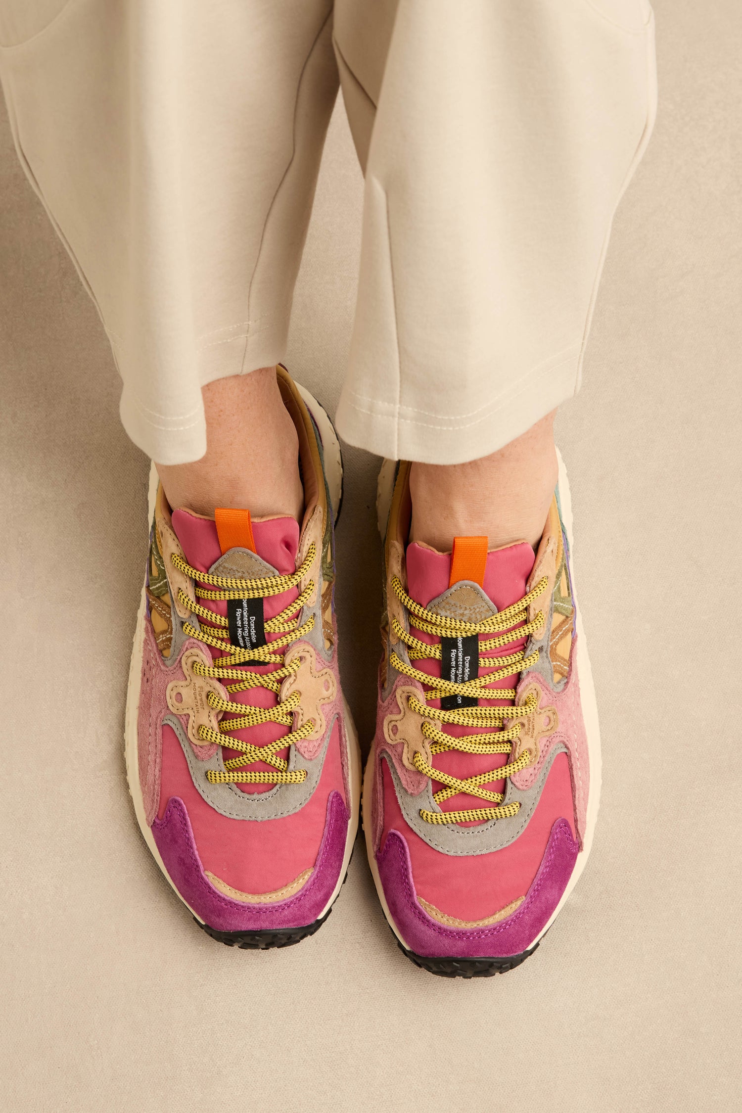 Top view of a person wearing light-colored pants and the Solin Colour Pop Trainers by Flower Mountain, crafted from suede and technical fabric with pink, purple, and yellow accents and multicolored laces—these vibrant trainers stand out beautifully against the beige surface.