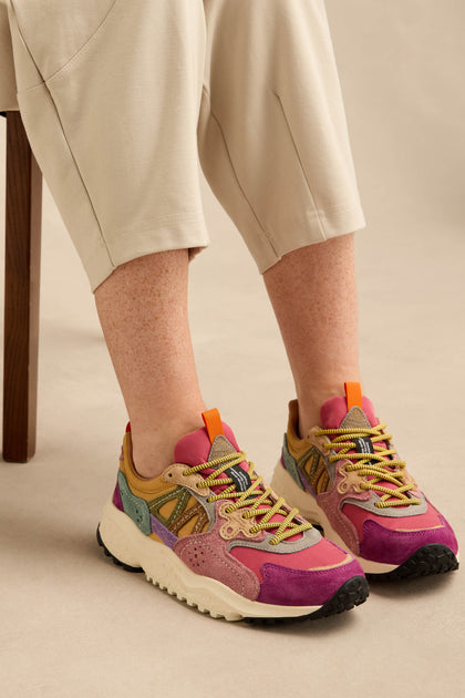 A person wearing beige pants and Solin Colour Pop trainers—crafted from suede and technical fabric with vibrant purple, green, tan, and orange accents—seated on a wooden chair against a neutral background.