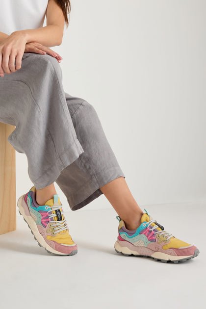 A person in loose grey pants and vibrant Dijon Yamano 3 Colour Pop Trainers sits on a light wooden stool against a plain white background.