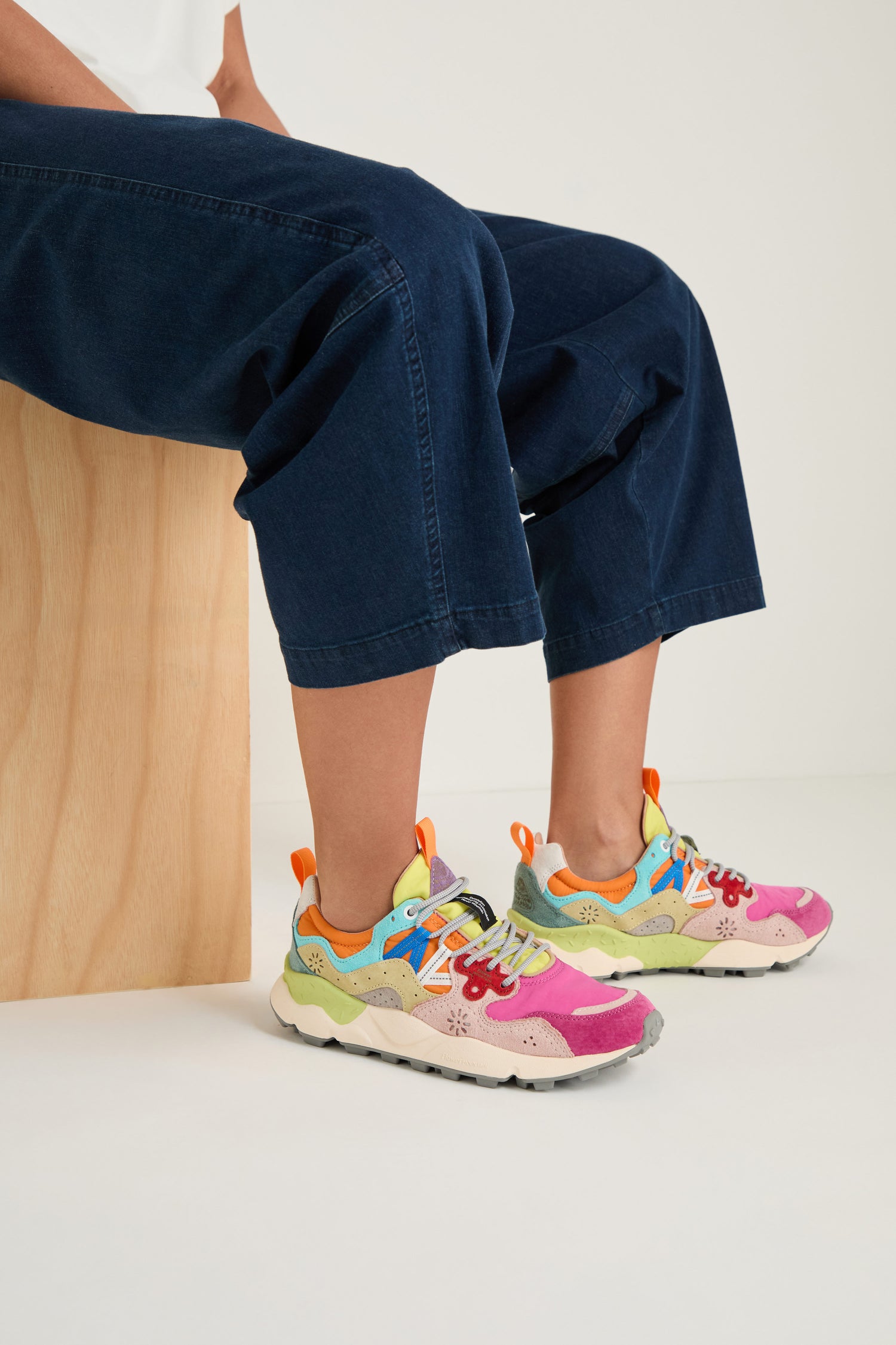 Person sitting on a wooden bench wears dark blue jeans and Fuchsia Yamano 3 Colour Pop Trainers—vibrant sneakers featuring pink, yellow, green, blue, and orange accents inspired by the outdoors.