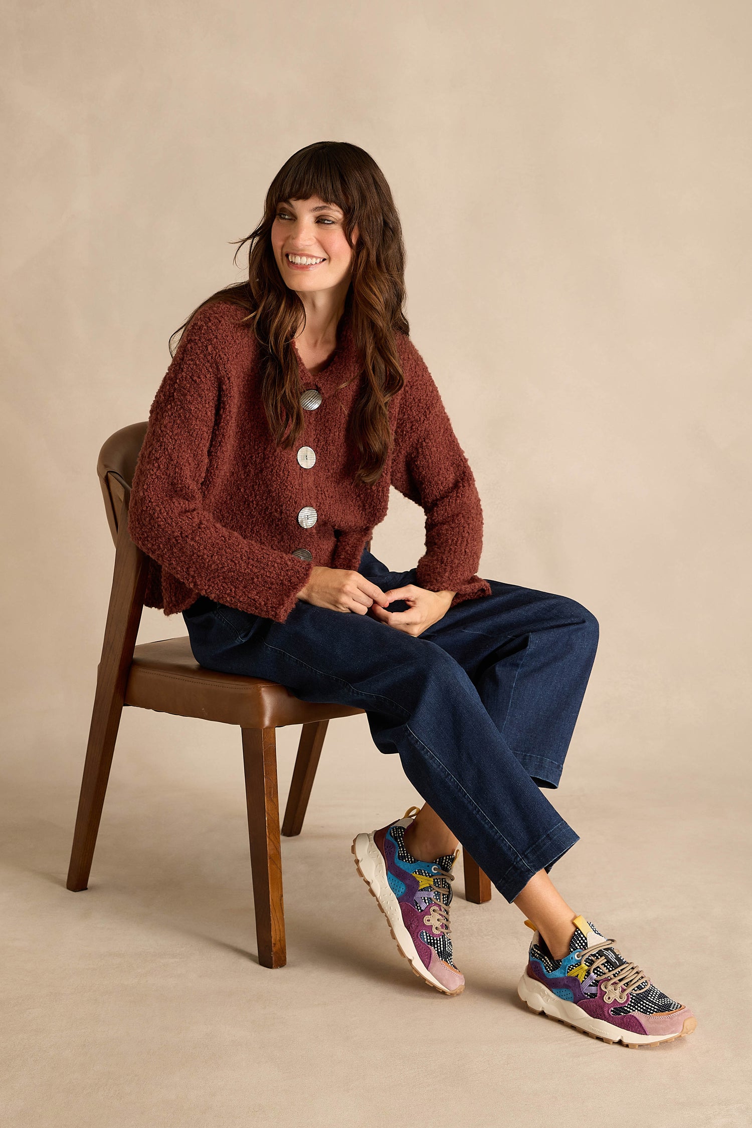 A woman seated on a brown chair wears a textured maroon cardigan, dark blue pants, and Yamano 3 Katsuo Sashi Trainers, posing against a plain beige background.