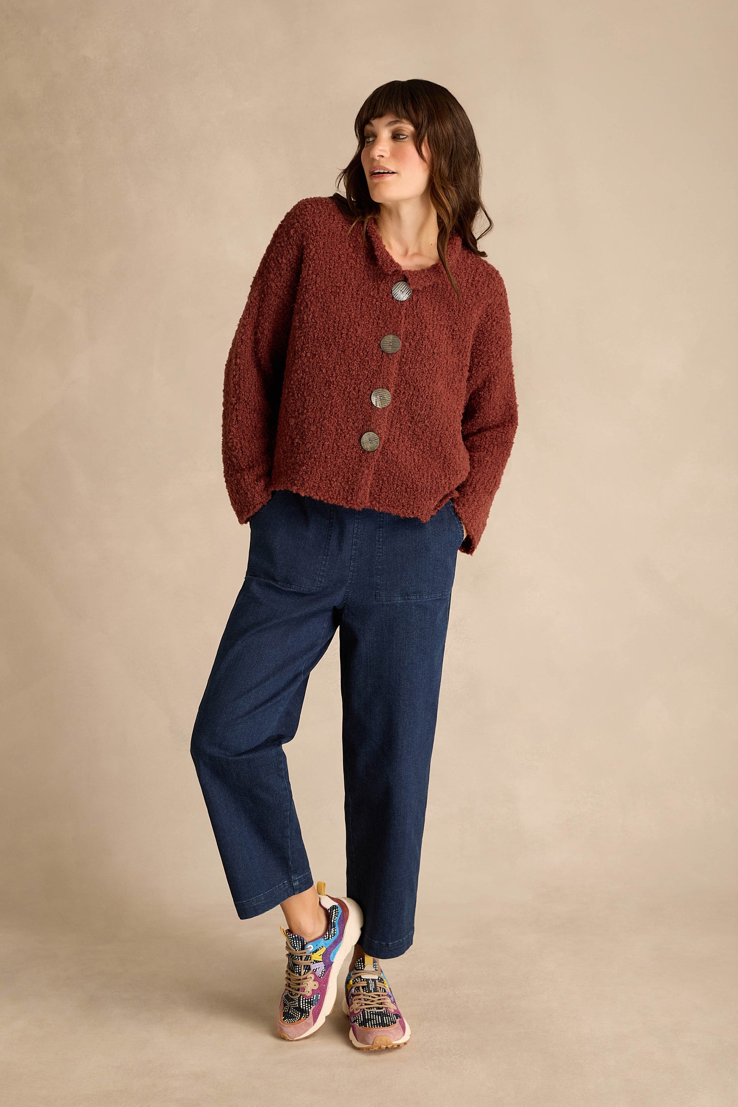 A woman stands against a plain beige background wearing a textured maroon button-up jacket, dark blue pants, and Yamano 3 Katsuo Sashi Trainers with trekking laces.