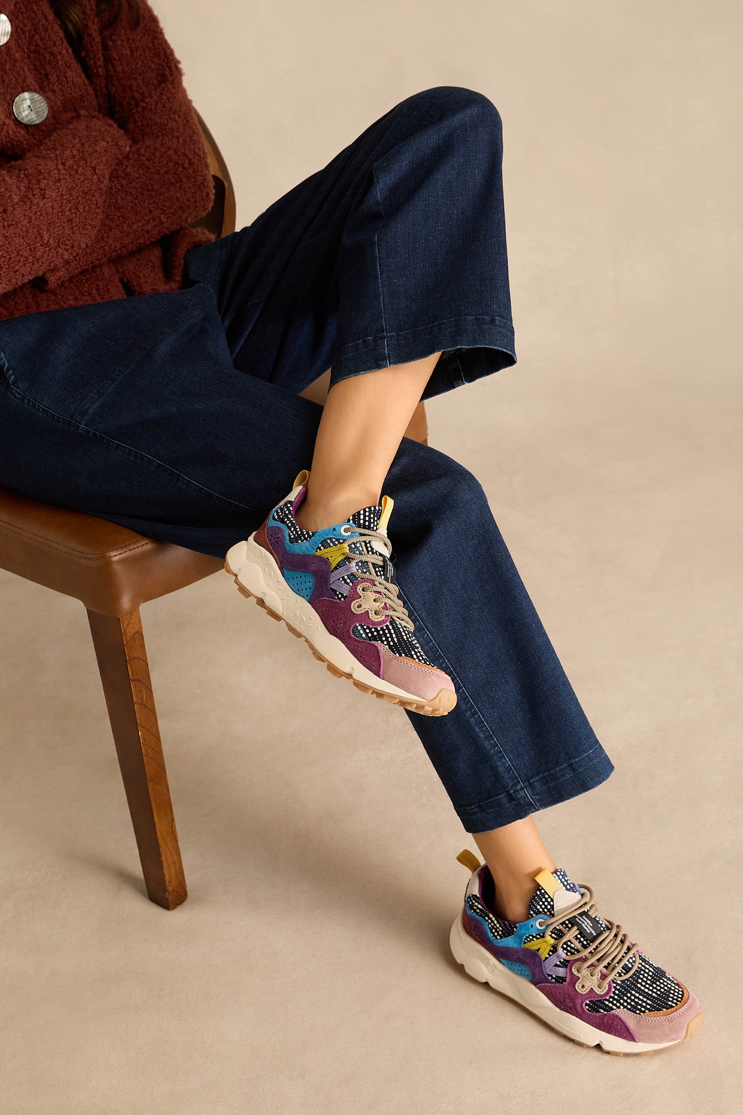 A person sits on a wooden chair wearing dark blue wide-leg jeans, a maroon fuzzy top, and vibrant Yamano 3 Katsuo Sashi Trainers—multi-colored patterned athletic shoes with chunky light soles.