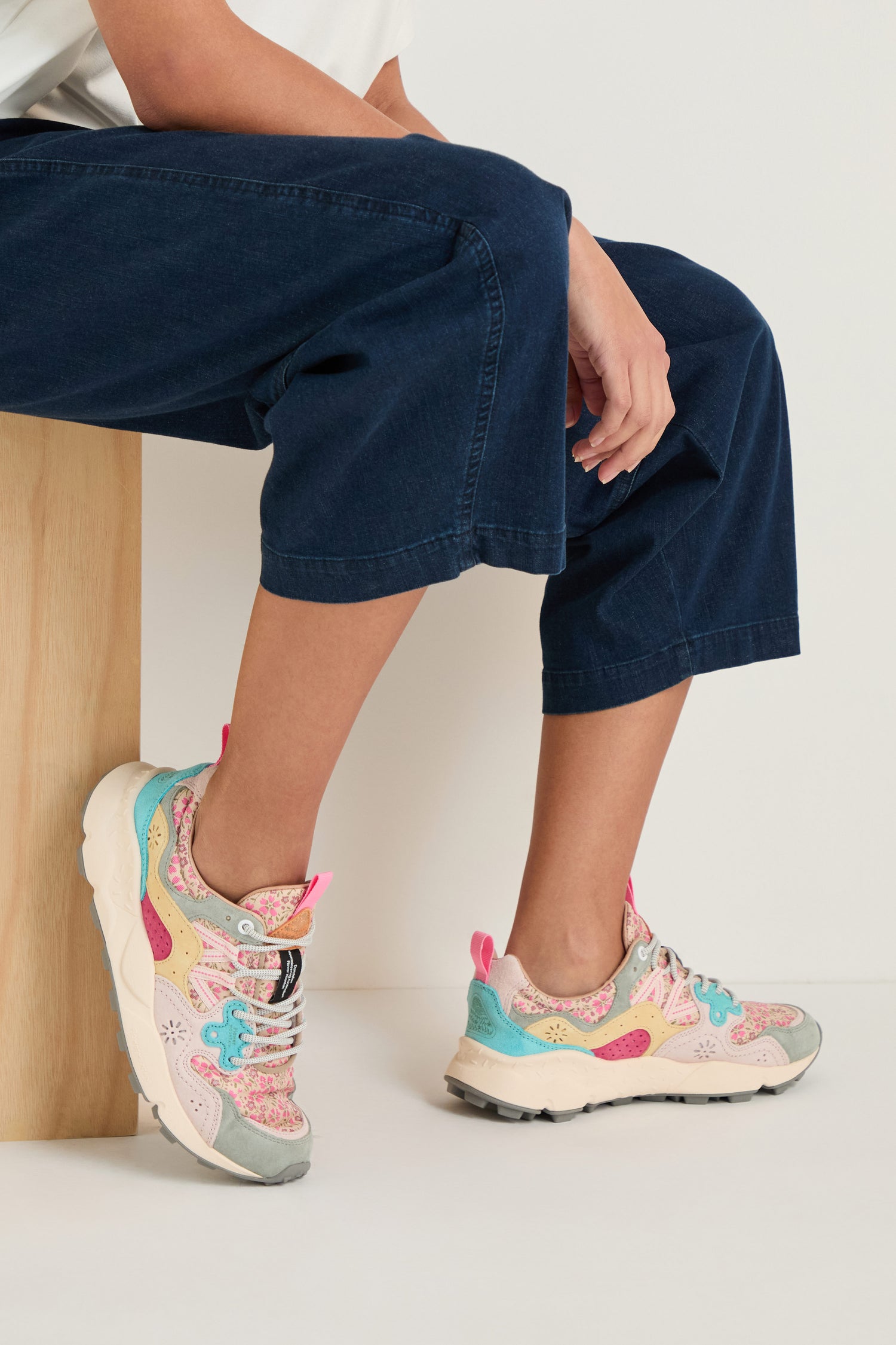 A person in dark blue pants and Yamano 3 Floral Trainers sits on a wooden bench with one leg crossed over the other.