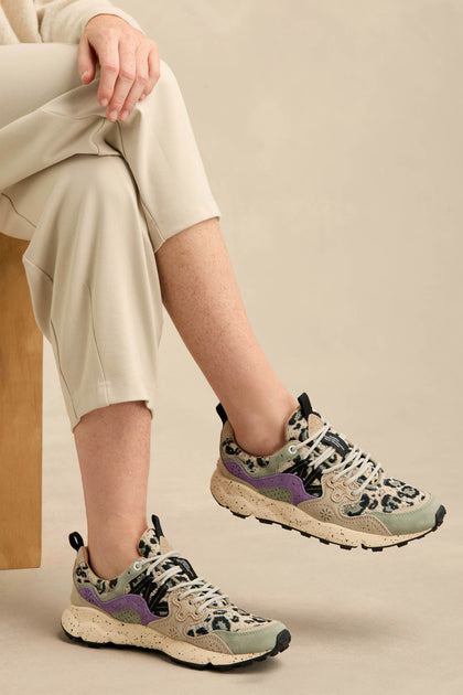 Person seated with legs crossed, wearing light-colored pants and Yamano 3 Leopard Print Trainers—beige chunky sole sneakers featuring black and purple accents and a leopard print pattern.