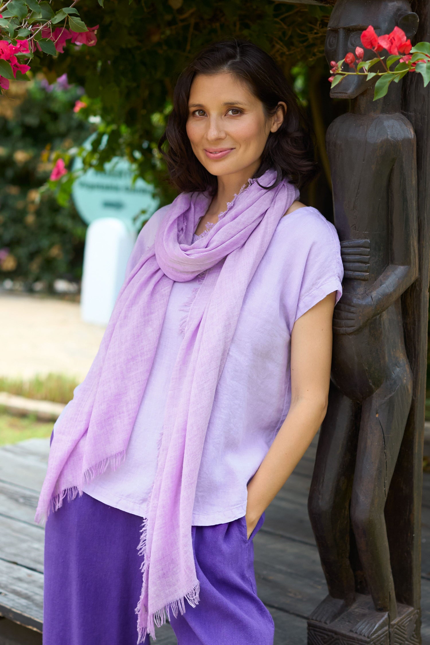 A woman in a purple outfit with a textured material Berry Bliss Scarf, smiling softly by a wooden statue and flowers.