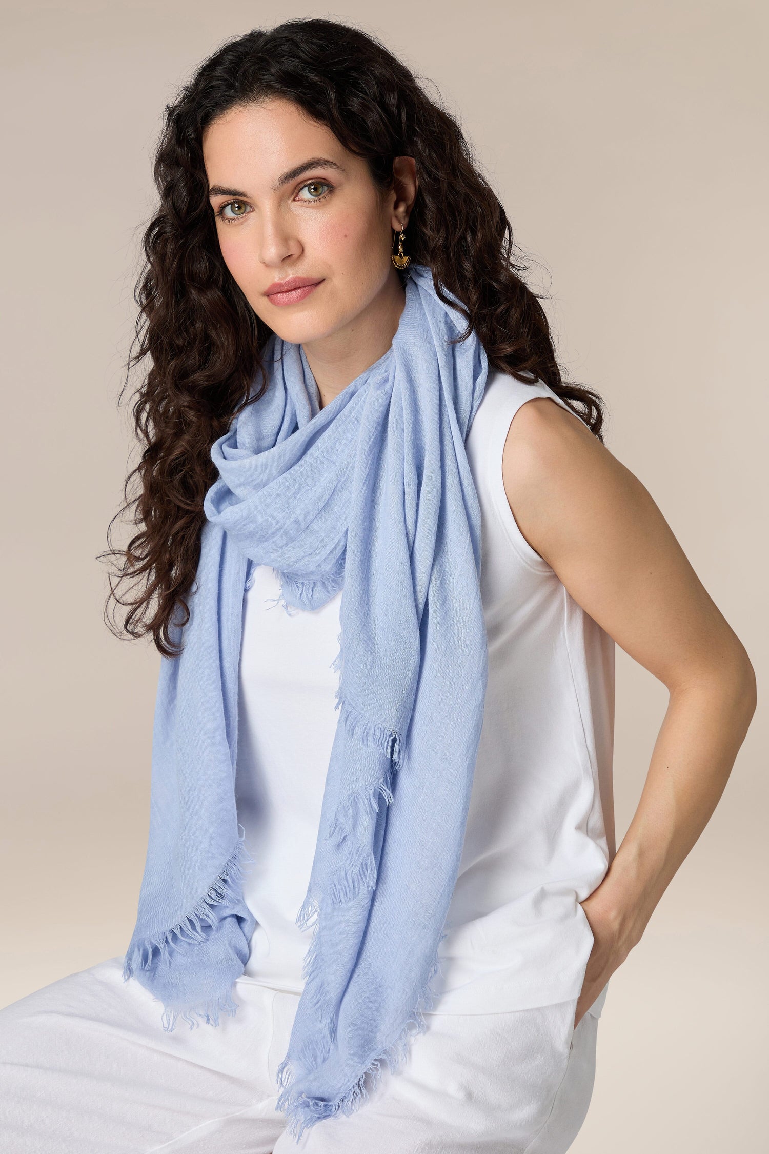 A woman with curly hair wearing a CozyMood Soft Fringe Scarf, white sleeveless top, and white pants.