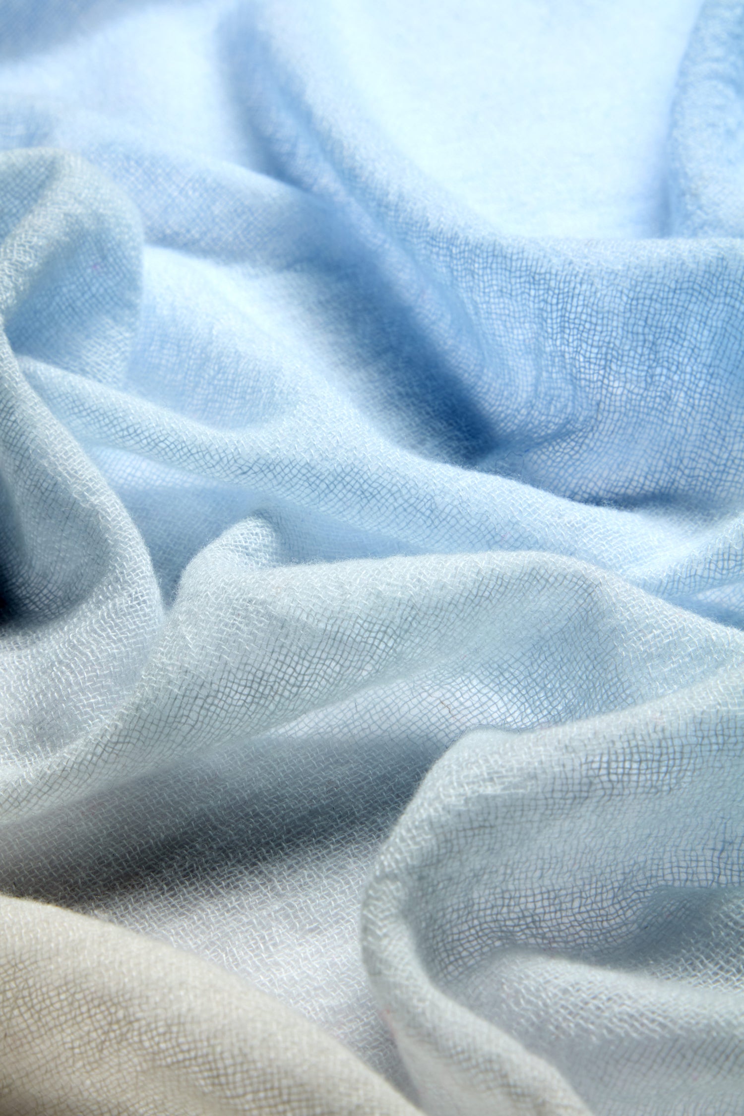 A close-up of soft, crumpled light blue fabric reveals the luxurious blend of the versatile Ombre Wool Scarf.