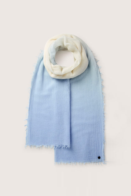 The Ombre Wool Scarf is a versatile accessory with a luxurious blend, showcasing a gradient design from white to light blue. It features fringed edges and is adorned with a small black button near the corner, making it perfect for any occasion.