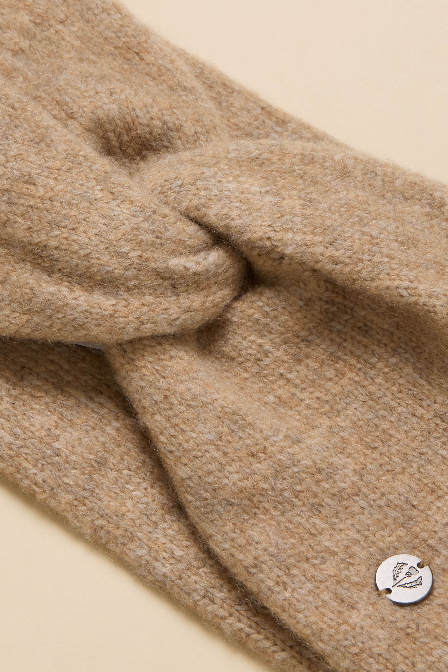 Close-up of the Wool Mix Sand Headband with a twisted knot detail and a small round metal logo tag, set against a beige background.