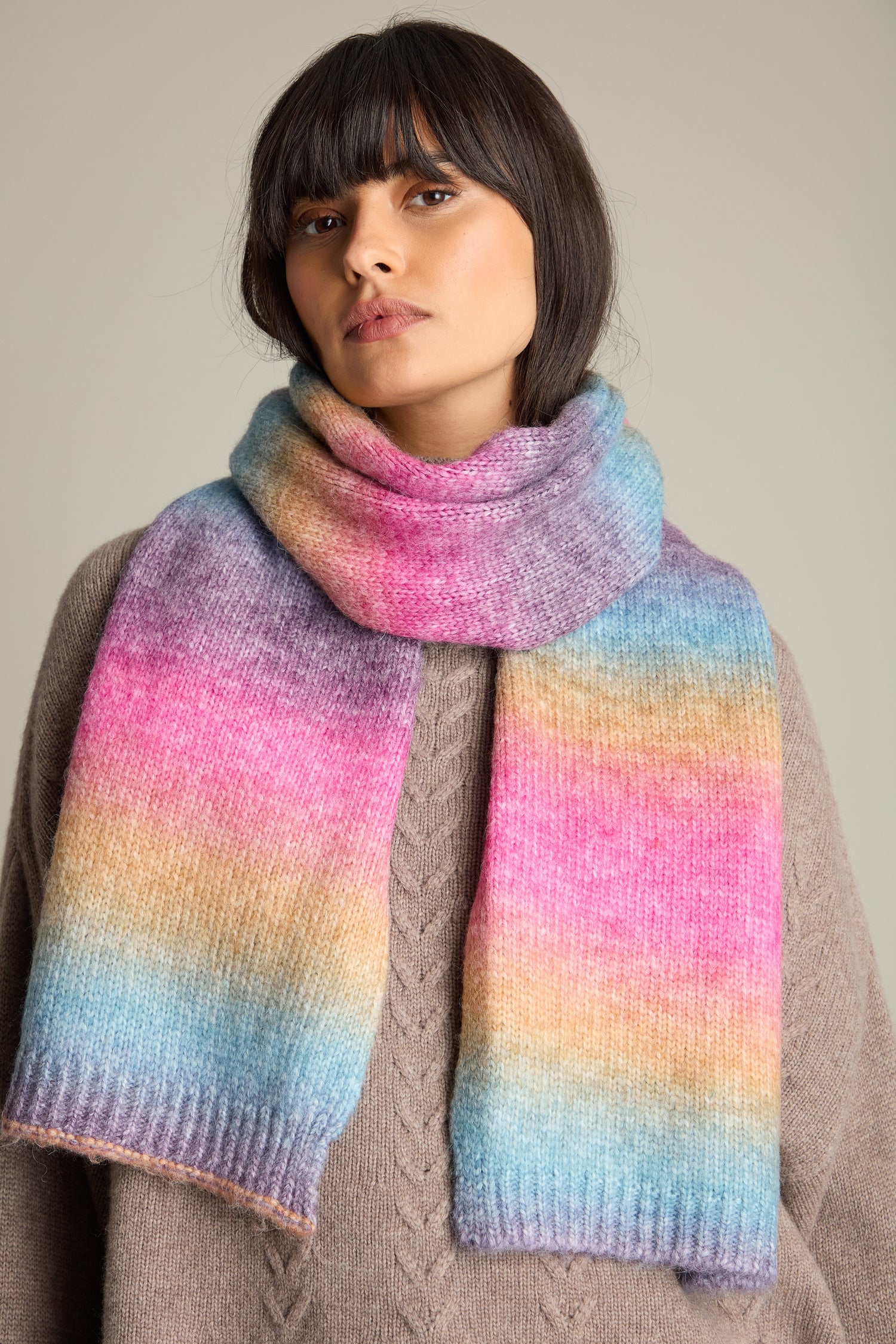 A person wearing a beige sweater and the Rainbow Stripe Knit Scarf featuring shades of pink, blue, and yellow stands against a neutral background.