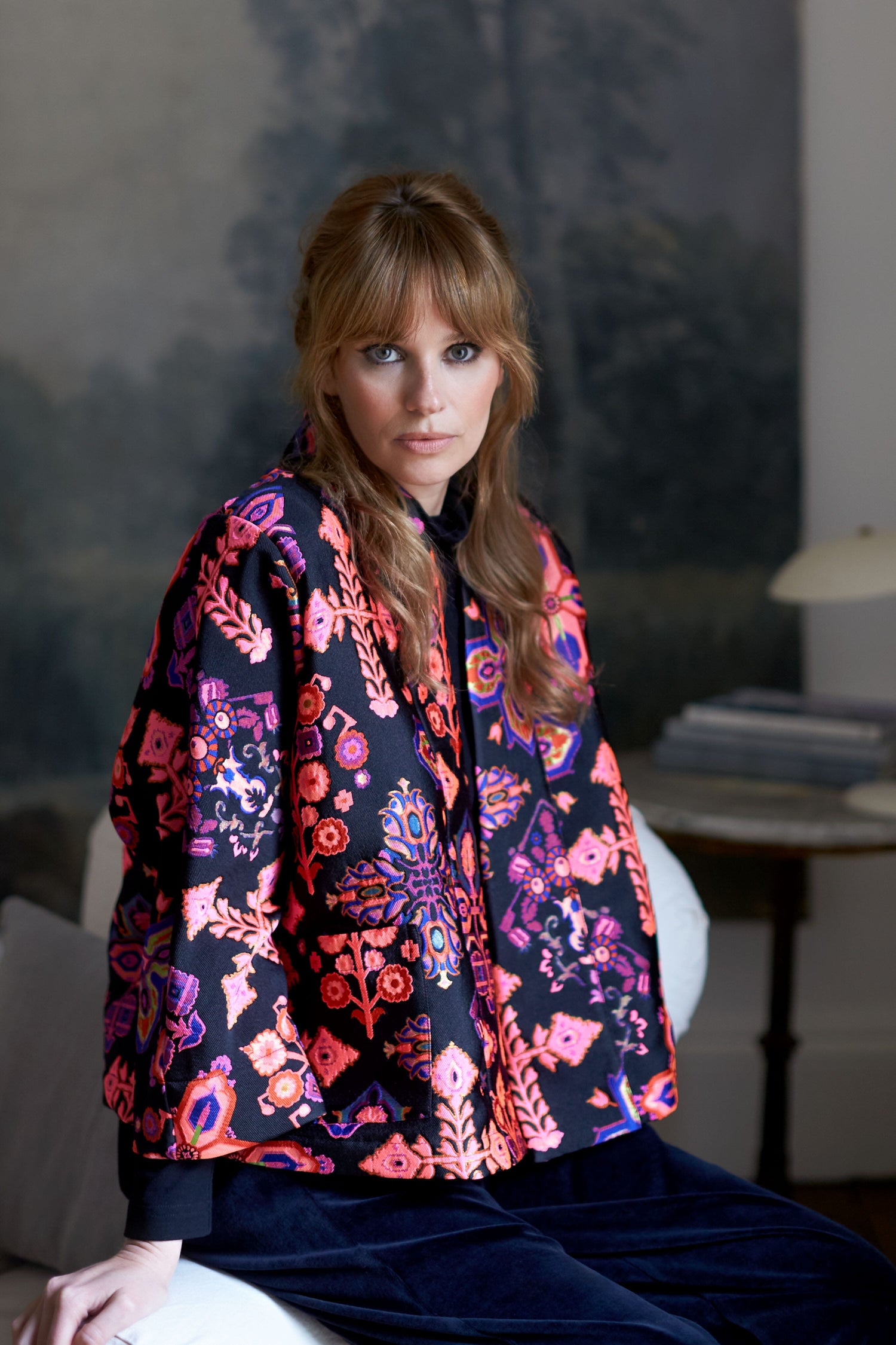 A woman is sitting on a chair in a room, wearing an exquisite Jacquard Aztec Floral Jacket made from jacquard fabric.