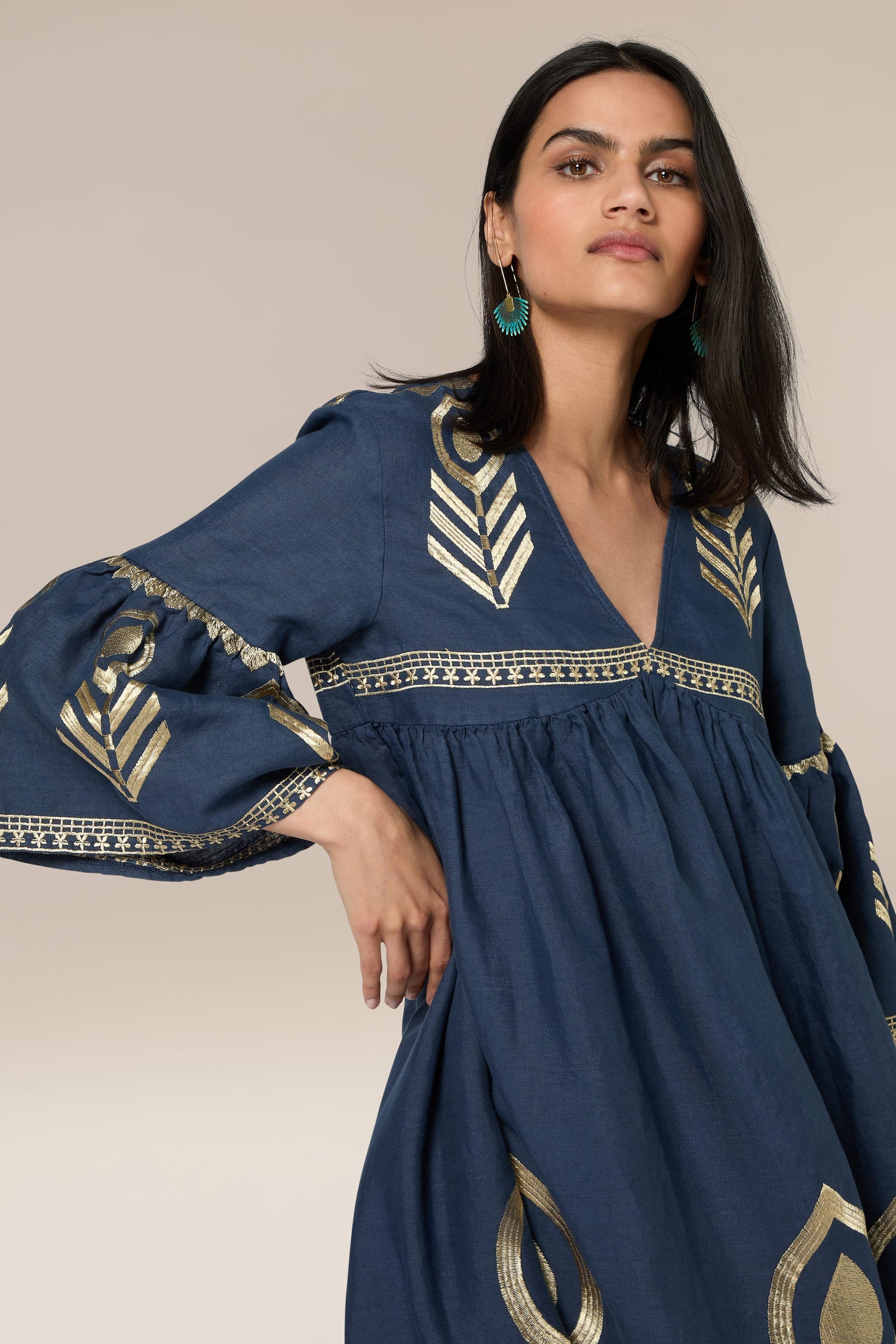 A woman stands wearing a Feathers Linen Midi Dress with gold patterns reminiscent of Greek Archaic Kori. She has long dark hair and is wearing green earrings. The background is plain beige.