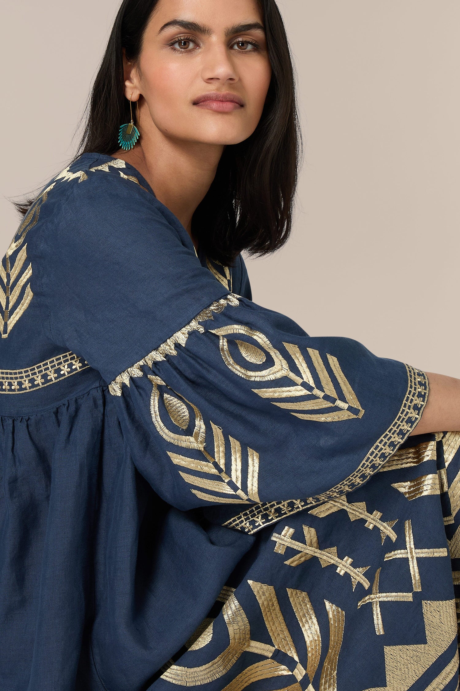 A woman is wearing a dark blue Feathers Linen Midi Dress featuring gold geometric patterns that echo Greek Archaic Kori designs. She has shoulder-length dark hair and is accessorized with a pair of green earrings, giving her look a touch of bohemian embroidery. She is looking slightly past the camera.