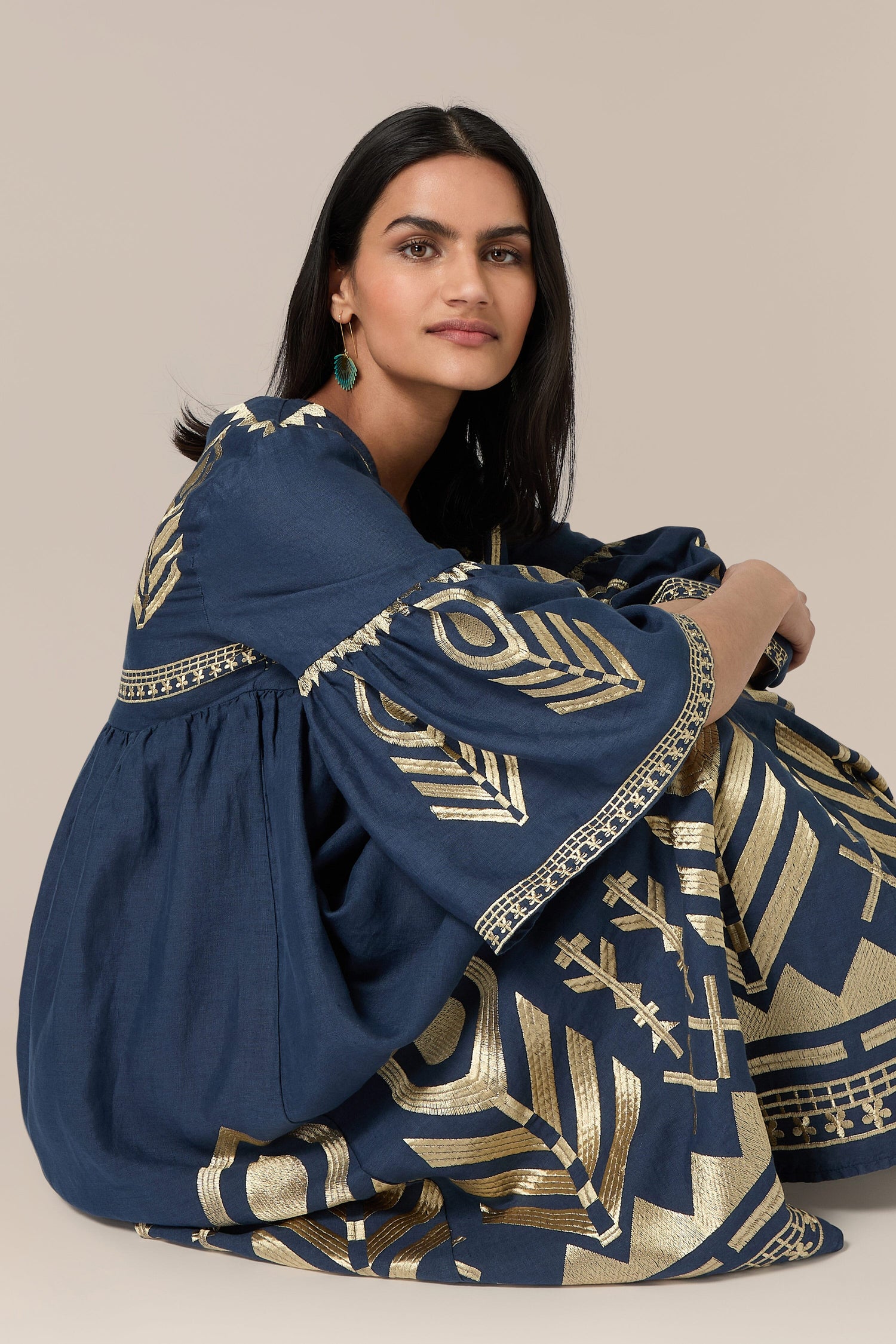 A person with long dark hair, wearing a Feathers Linen Midi Dress adorned with bohemian embroidery and gold geometric patterns, sits on the floor with legs crossed and looks into the camera against a beige background.