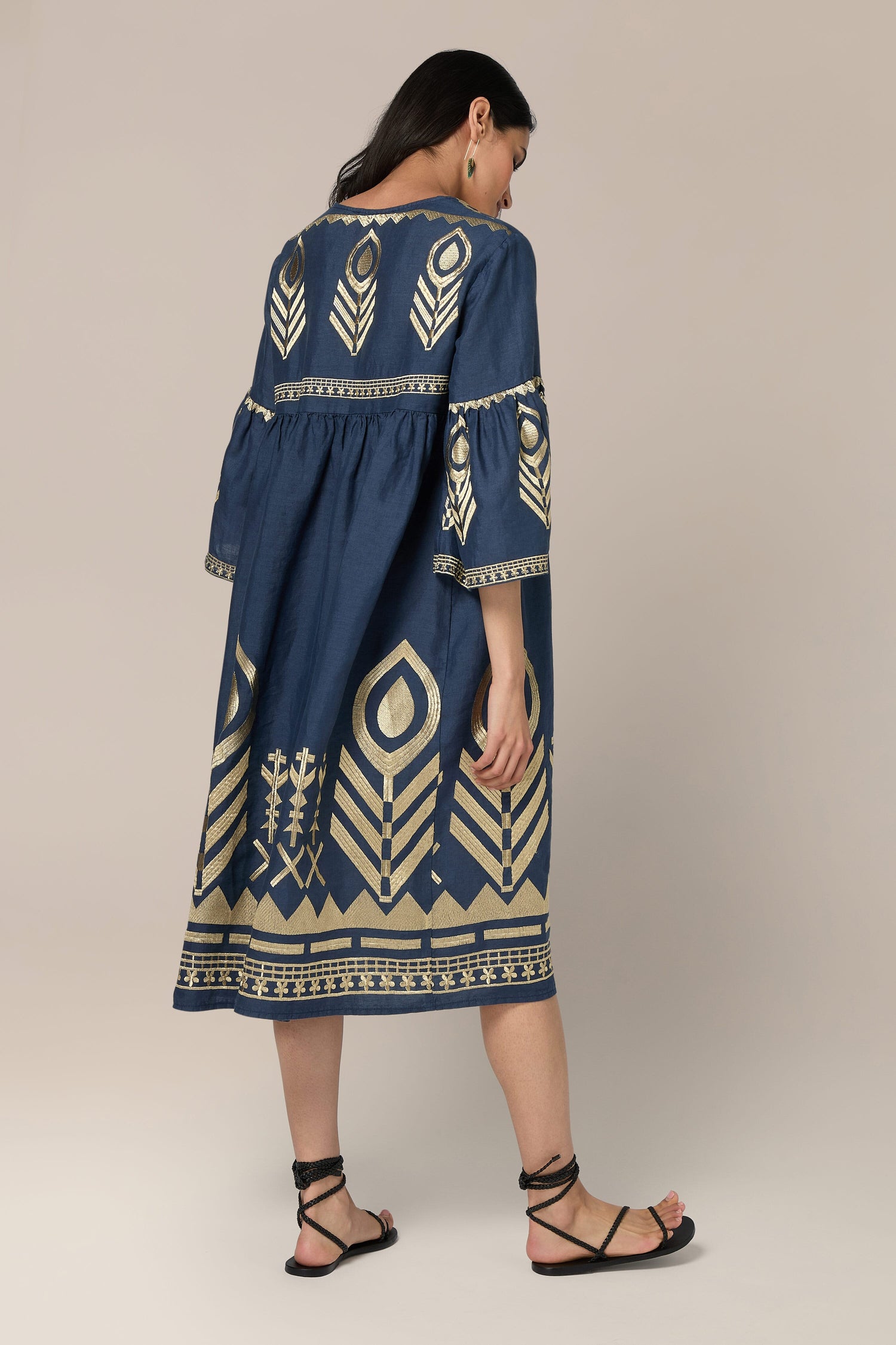 A woman is standing and facing away, wearing a Feathers Linen Midi Dress with intricate gold patterns reminiscent of Greek Archaic Kori. She has long dark hair and is wearing black strappy sandals.