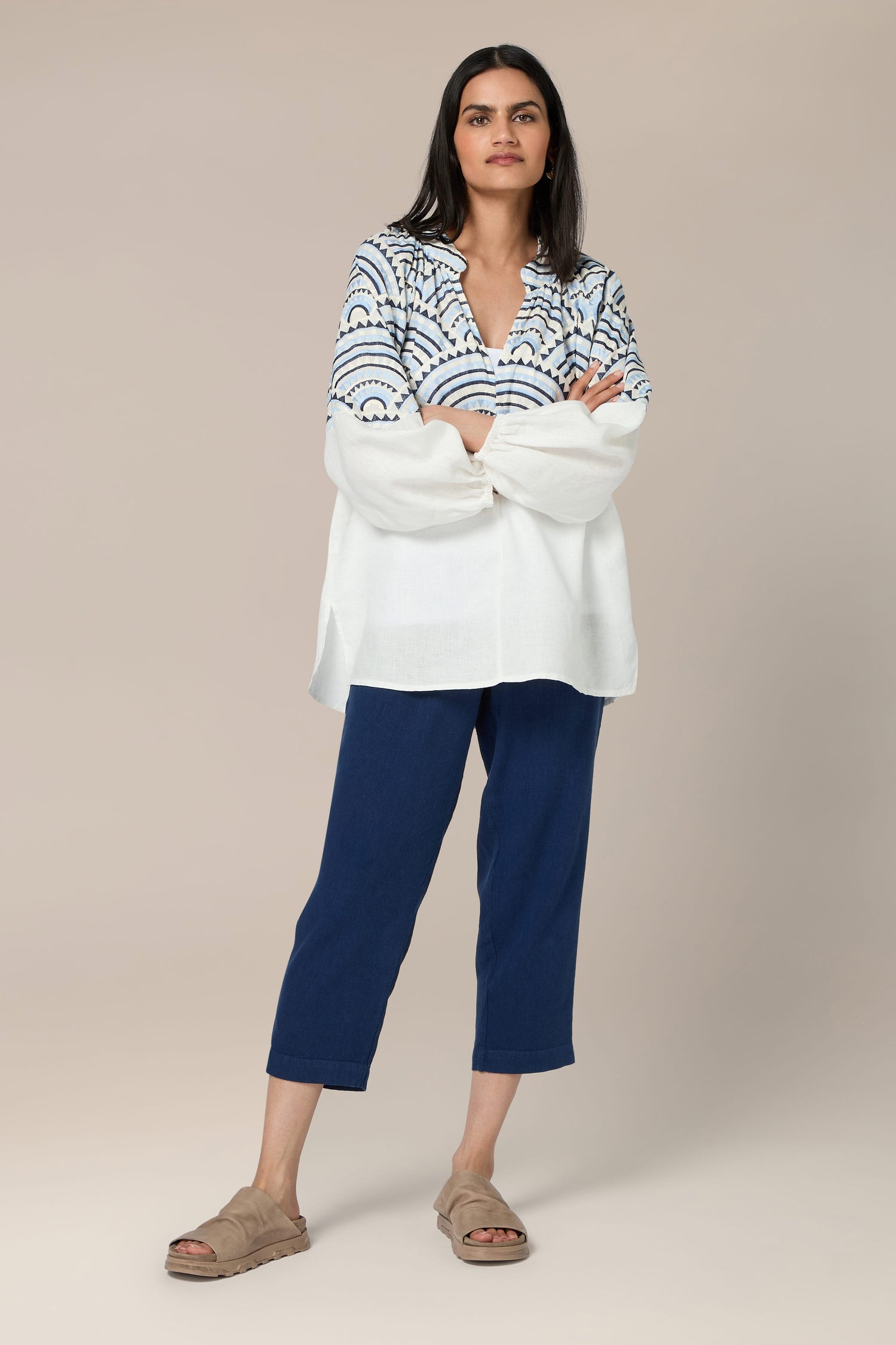 A person with long hair stands with arms crossed, wearing a Sundial Linen Blouse, blue pants, and beige sandals against a plain background.