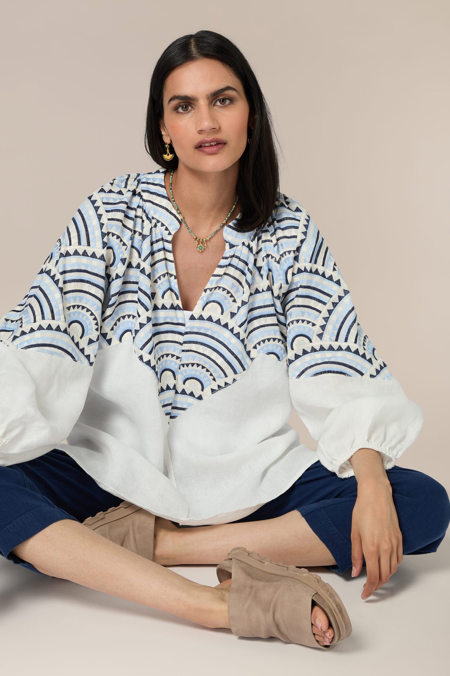 A woman with dark hair sits cross-legged, wearing a white and blue Sundial Linen Blouse adorned with intricate embroidery, blue pants, and beige slip-on shoes. The Sundial Linen Blouse features elegant balloon sleeves. She has a neutral expression and is looking at the camera.