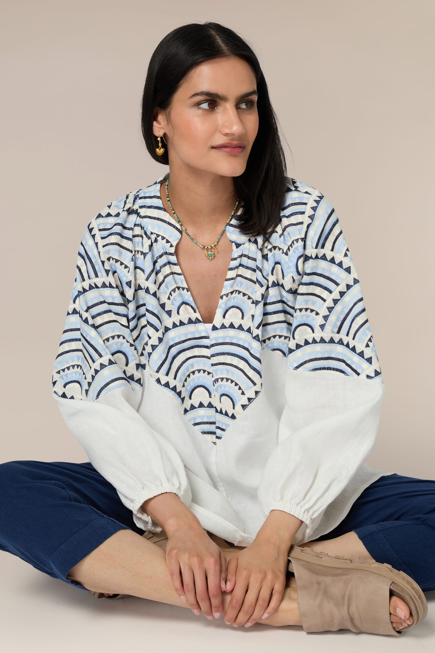 A seated woman with dark hair, wearing a Sundial Linen Blouse adorned with intricate embroidery and blue pants, looks to her left against a beige background.