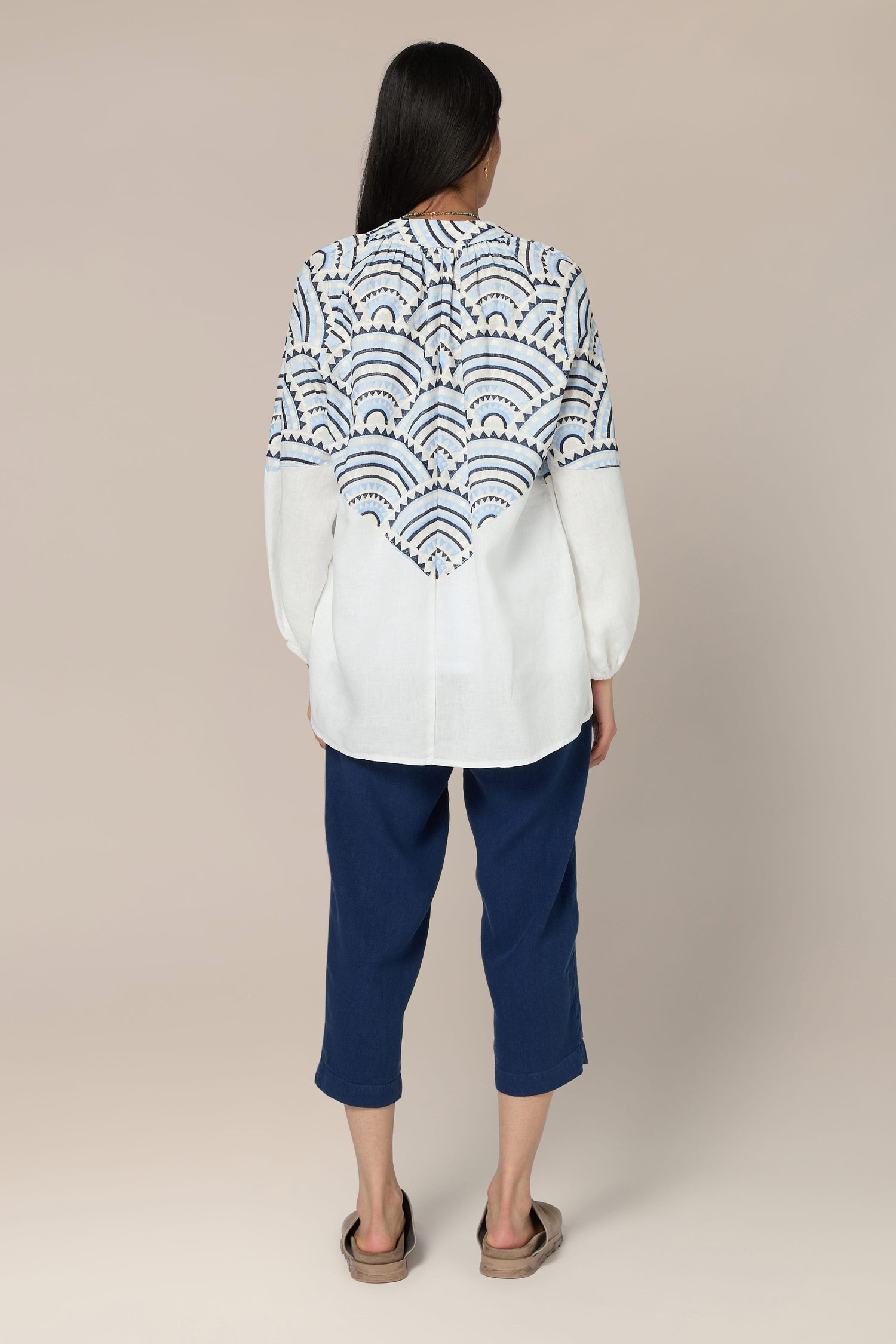 A person with long hair is standing, facing away. They are wearing a white Sundial Linen Blouse with intricate embroidery on the back, blue cropped pants, and brown sandals.