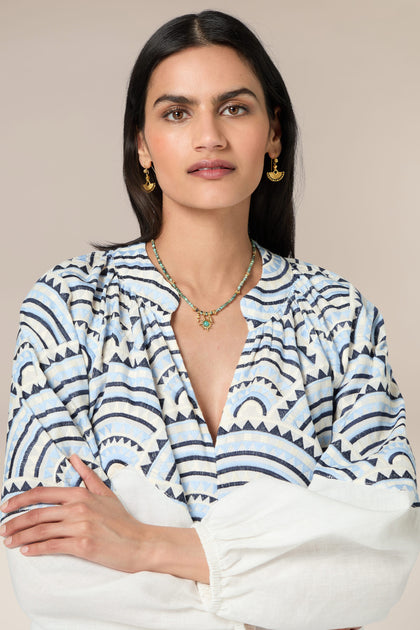 A woman with long dark hair wears a Sundial Linen Blouse, gold earrings, and a necklace. She stands with her arms crossed against a neutral background.