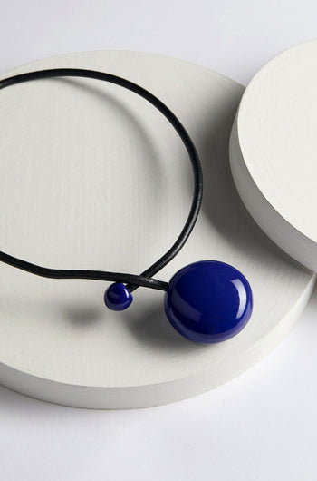 The Handmade Ceramic Bead Necklace features a large circular blue charm and a smaller blue bead on a black cord, exemplifying French craftsmanship and contemporary design, beautifully displayed against a white round platform background.