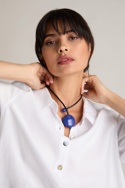 A person wearing a white blouse adjusts a black Handmade Ceramic Bead Necklace, which features a large blue pendant and showcases the elegance of contemporary design.