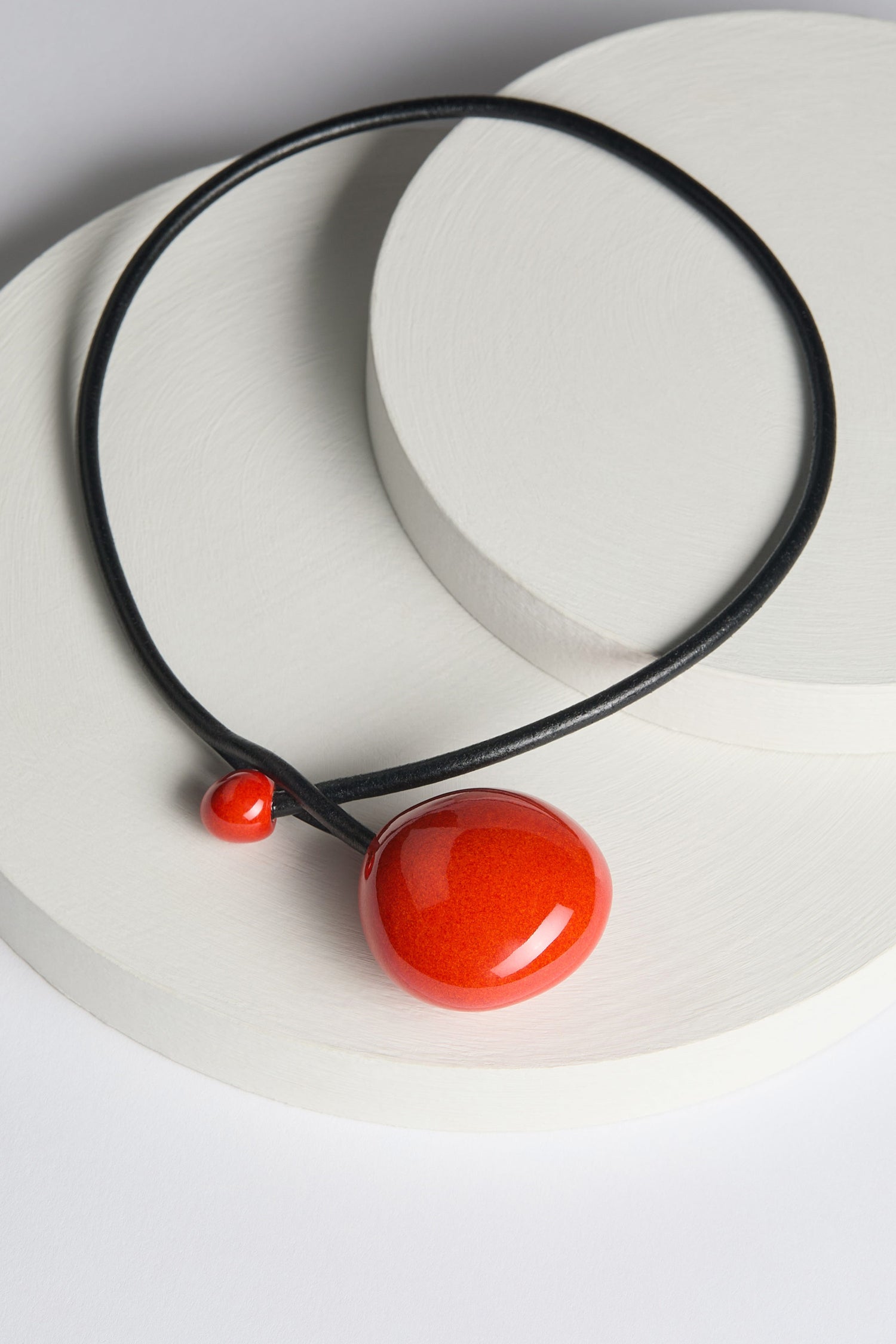 The Handmade Ceramic Bead Necklace features a minimalist black design adorned with a large red circular pendant and a small red bead, all elegantly displayed on a round white ceramic platform, exuding contemporary aesthetics and exquisite French craftsmanship.