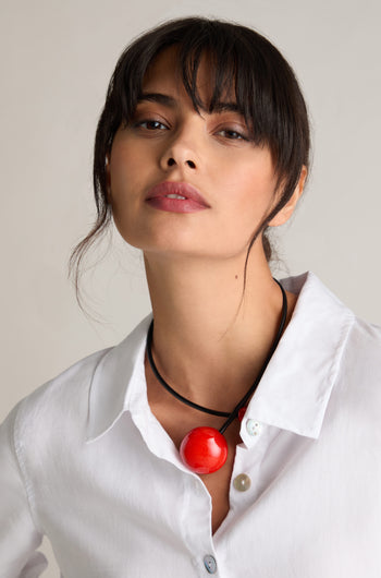 A person with dark hair wears a white shirt and the Handmade Ceramic Bead Necklace with a large red pendant, showcasing contemporary design accented by French craftsmanship.