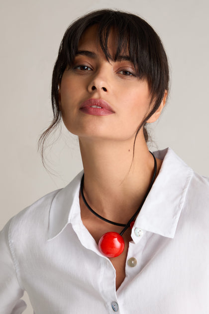 A person with dark hair wears a white shirt and the Handmade Ceramic Bead Necklace with a large red pendant, showcasing contemporary design accented by French craftsmanship.