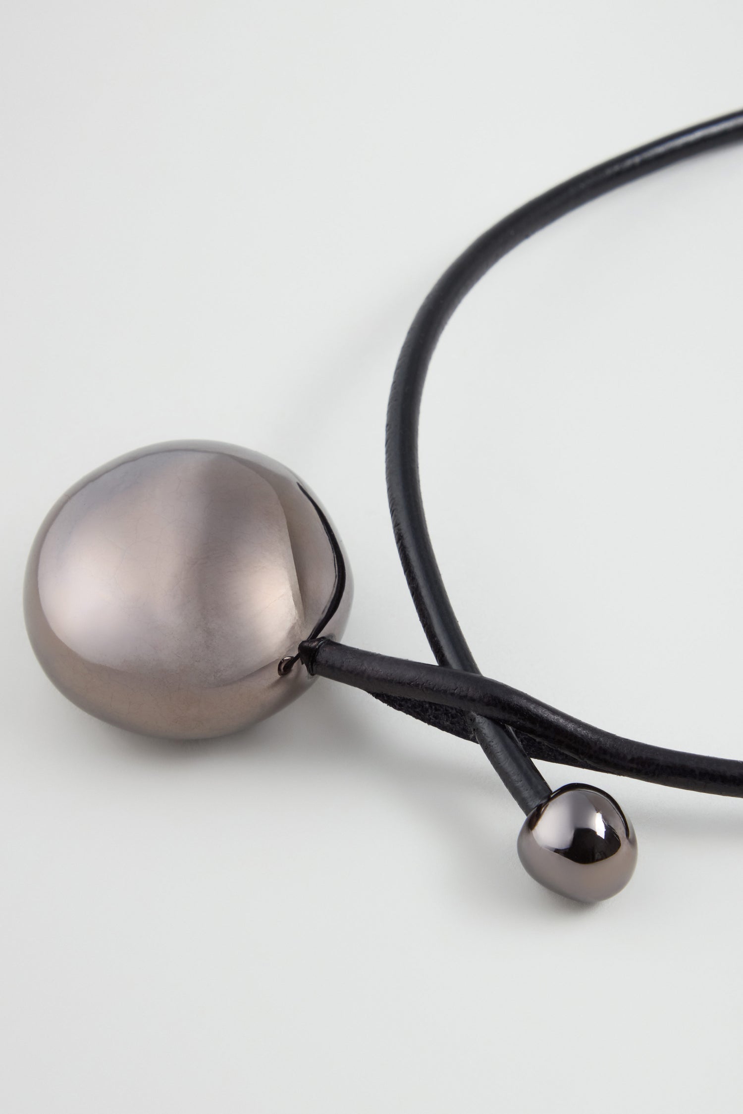 The Charcoal Handmade Ceramic Bead Necklace features a black cord with two metallic spherical pendants, one large and one small, highlighting exquisite French design against a white background.