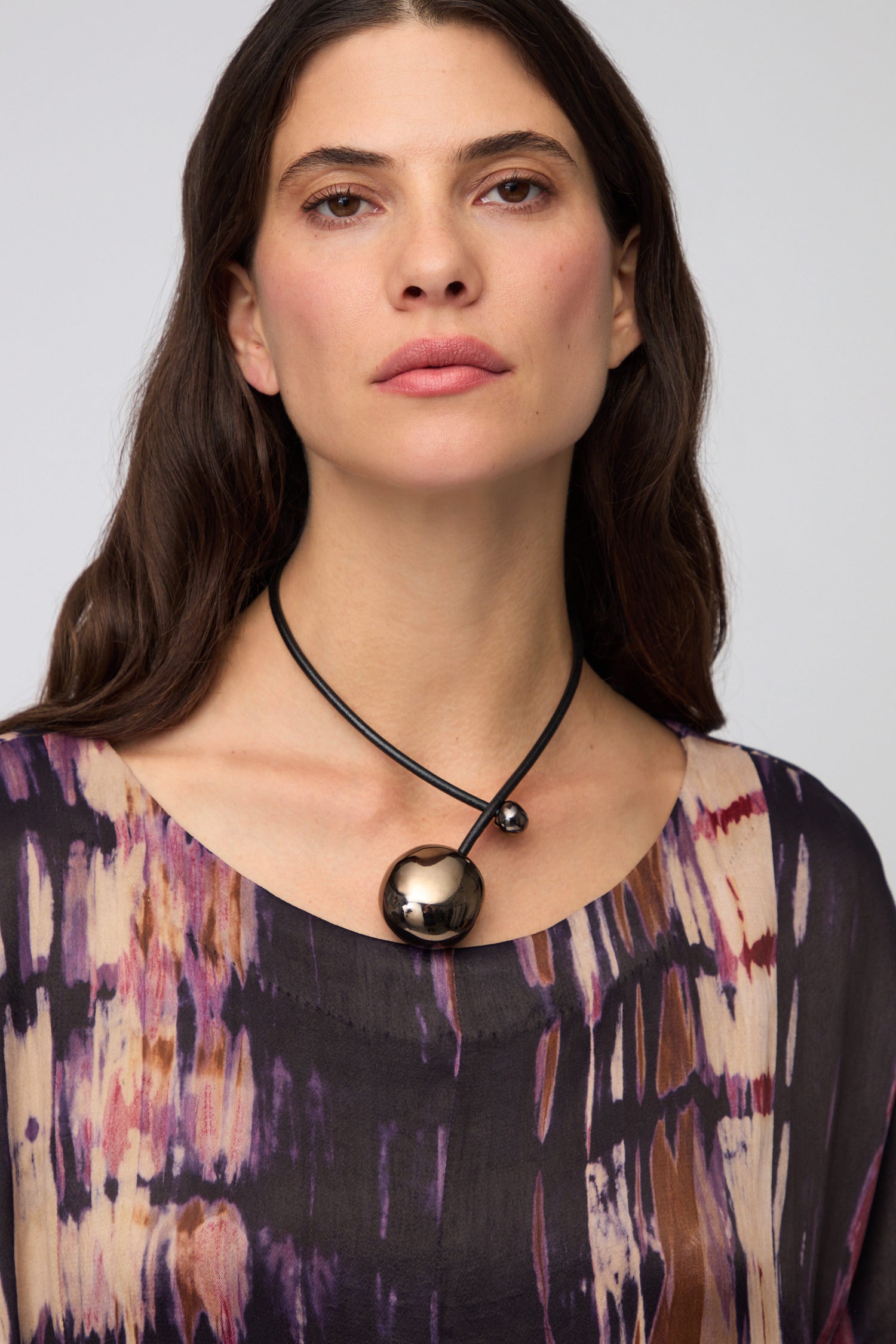 A woman with long dark hair and a patterned top wears the Charcoal Handmade Ceramic Bead Necklace—an artisan piece featuring two shiny ceramic beads on a black cord. She gazes directly at the camera with a neutral expression.
