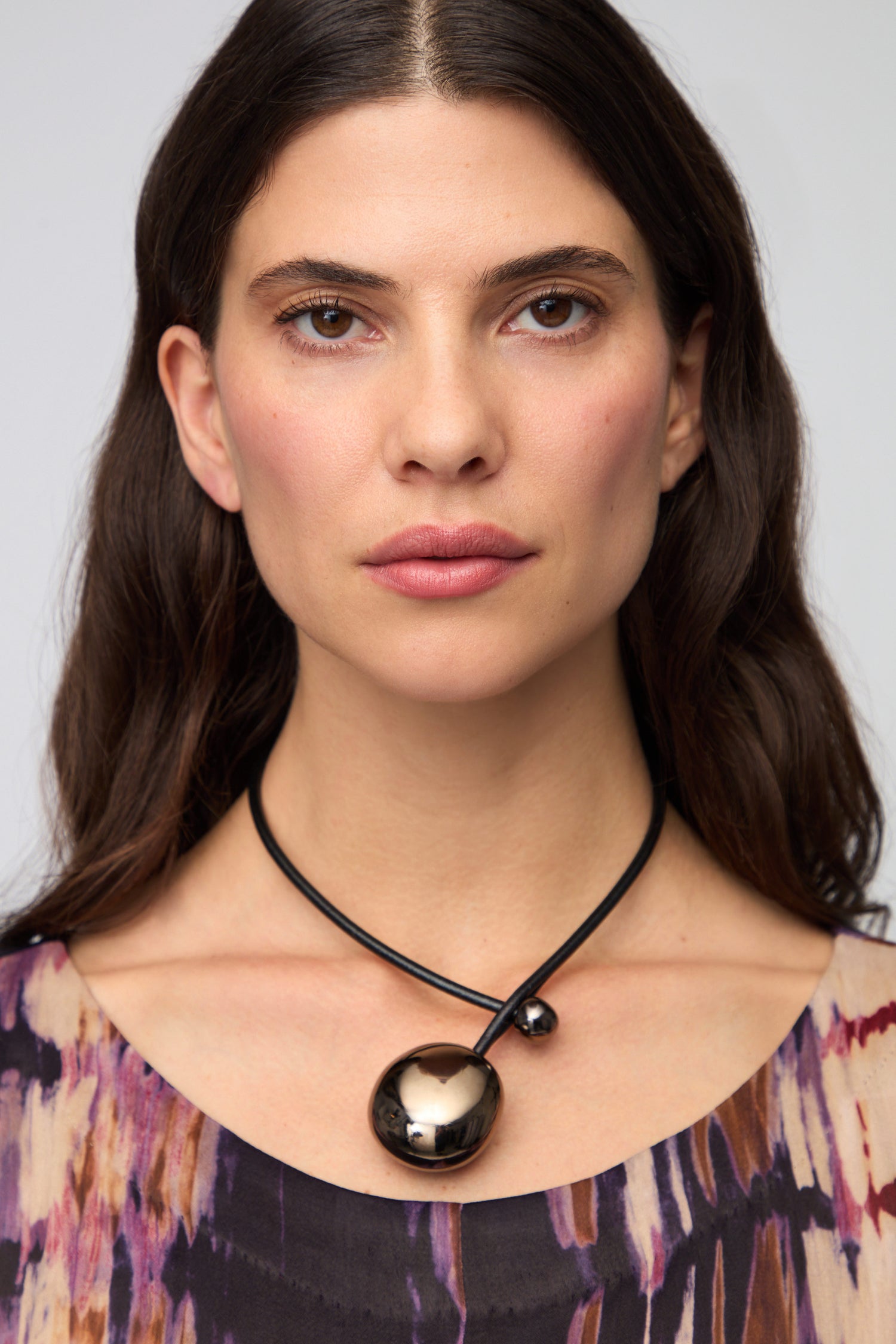 A woman with long dark hair in a patterned top wears the Charcoal Handmade Ceramic Bead Necklace—featuring a black cord and large ceramic bead—facing forward against a plain background.