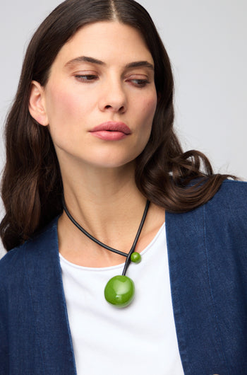 Woman with long brown hair wearing a blue jacket, white shirt, and the Moss Handmade Ceramic Bead Necklace featuring a large green pendant, looking to her left.