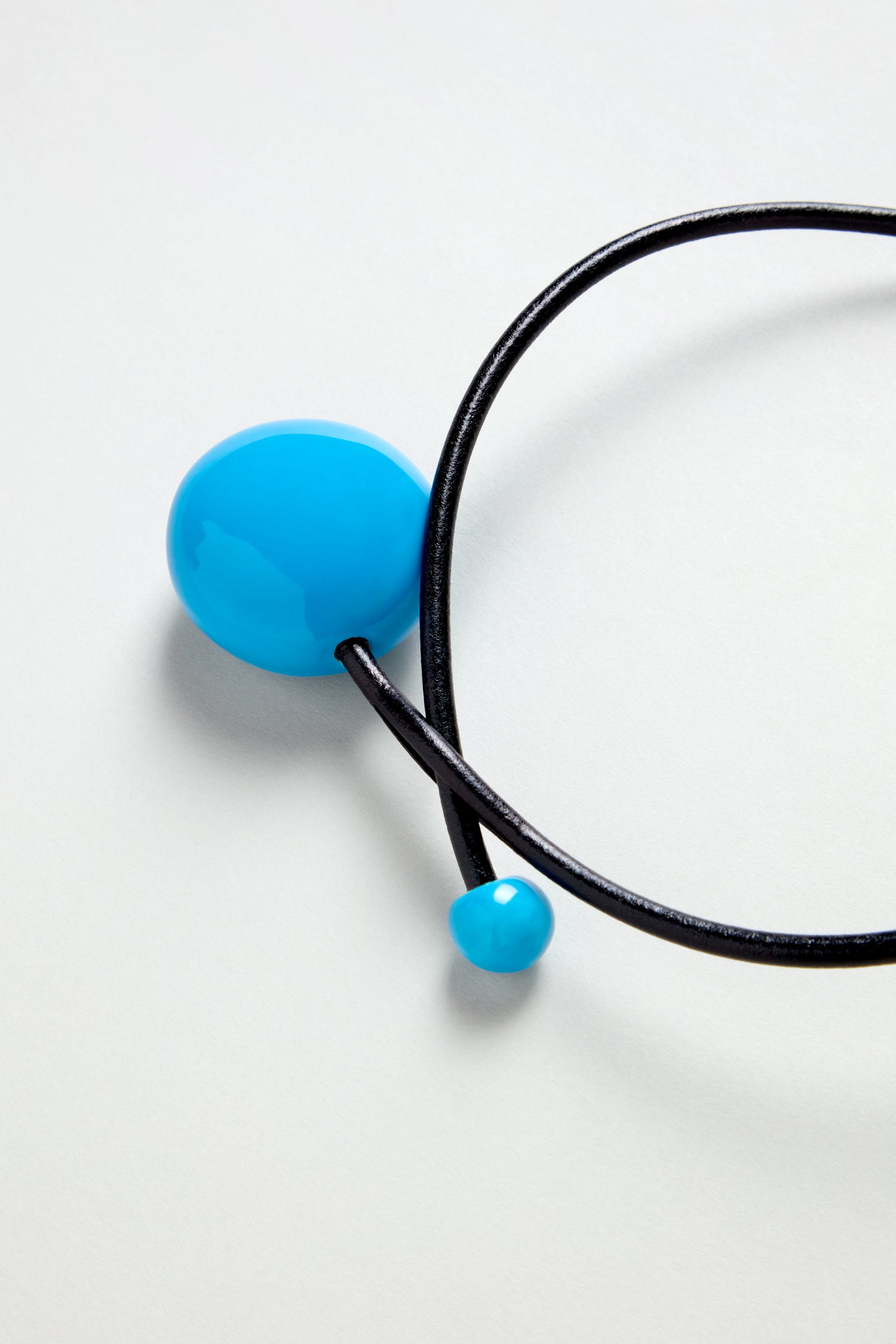 The Handmade Ceramic Bead Necklace features a black leather cord with two glossy blue ceramic beads, one large and one small, handcrafted in France and shown on a light gray background.