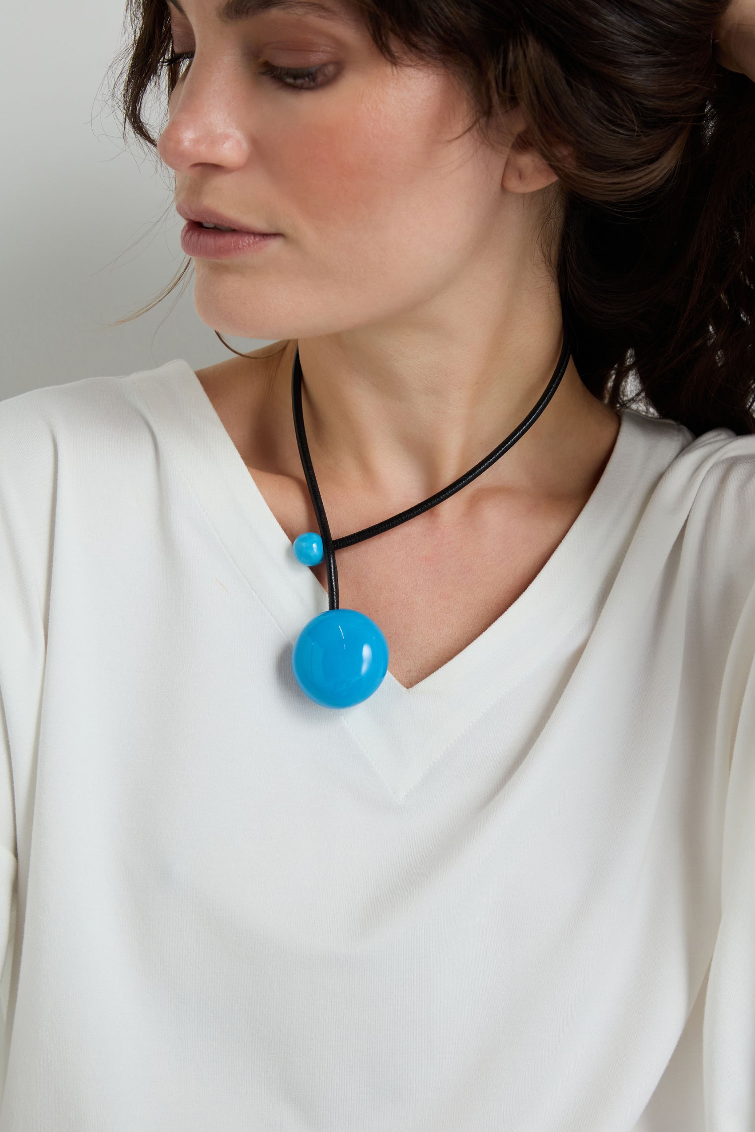 A person wears a white V-neck top and the Handmade Ceramic Bead Necklace—a black leather cord with two round blue ceramic beads, one large and one small—offering an elegant, artisanal look.