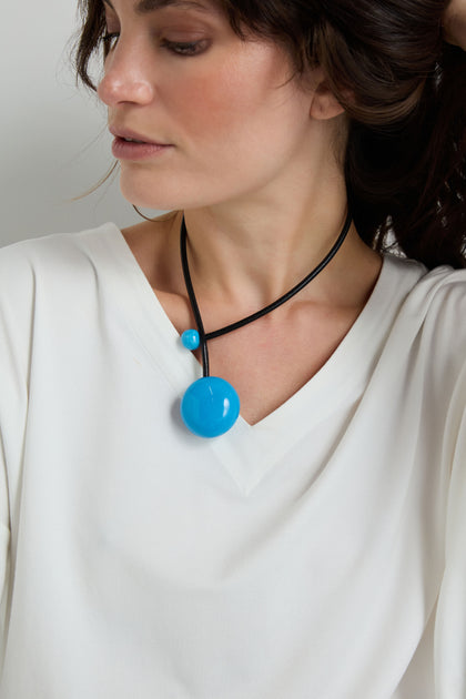 A person wears a white V-neck top and the Handmade Ceramic Bead Necklace—a black leather cord with two round blue ceramic beads, one large and one small—offering an elegant, artisanal look.