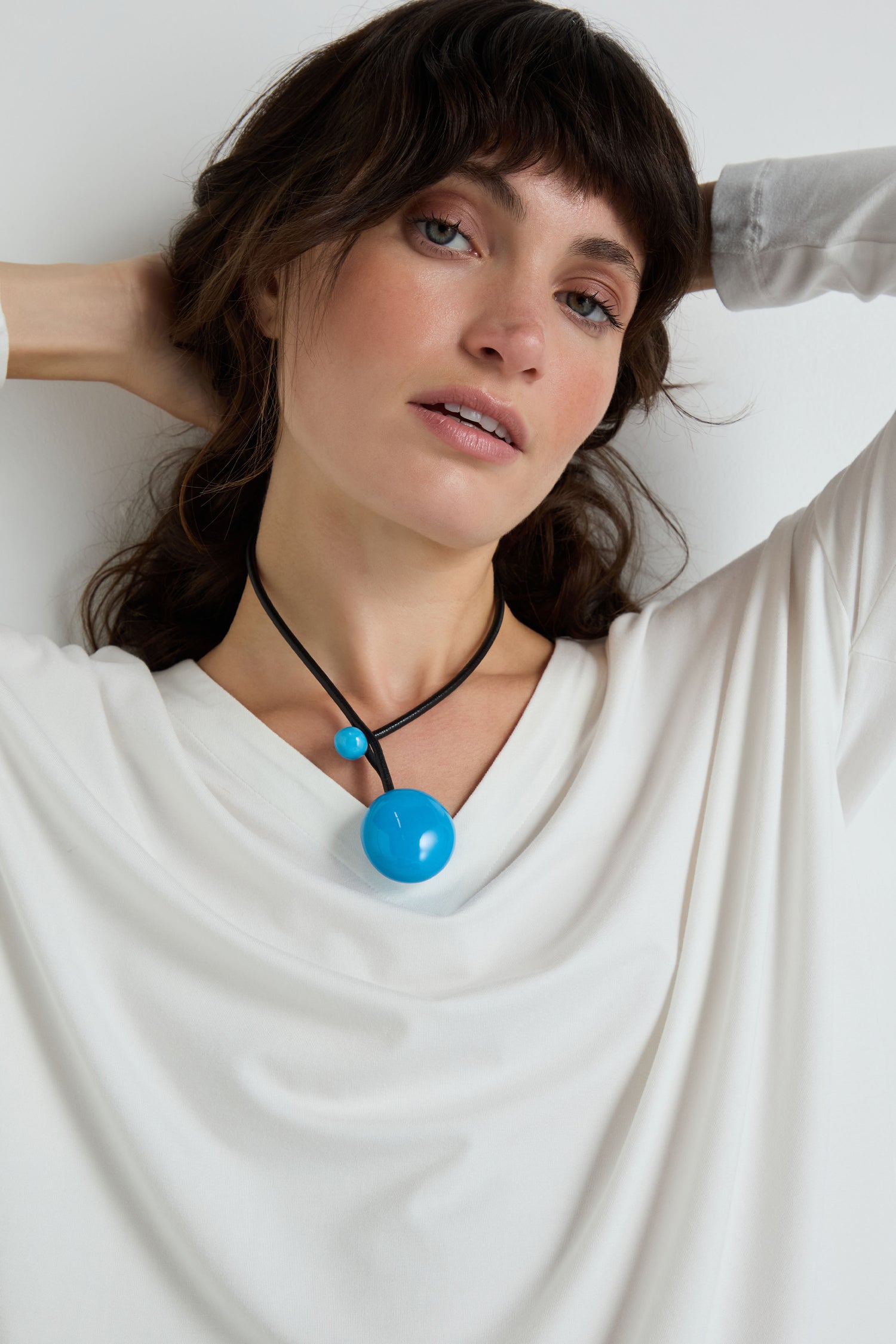 A woman in a white top poses with her hands behind her head, wearing the Handmade Ceramic Bead Necklace featuring two blue spherical pendants.
