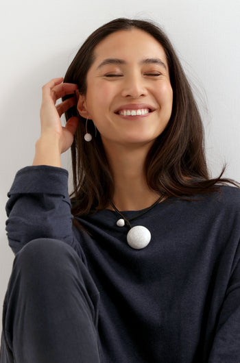 A woman with long dark hair smiles, wearing a dark top and the Handmade Ceramic Crackle Bead Necklace, with matching earrings, seated against a light background.